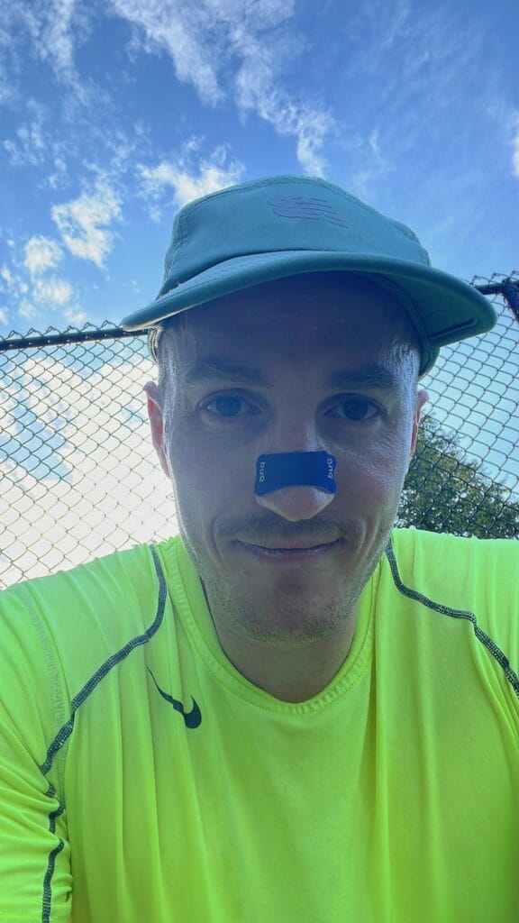 Man in a cap and neon shirt wearing a nasal strip, taking a selfie on a court