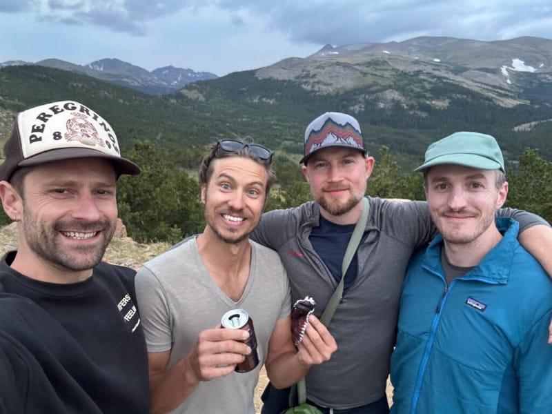 Four men smiling on a mountain overlook, holding drinks