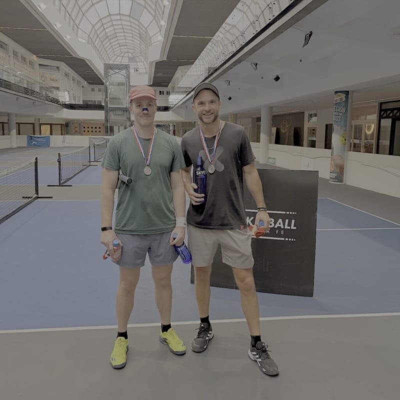 Two men wearing medals, one holding a trophy, on an indoor pickleball court